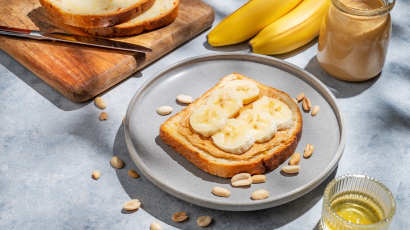 Tost sa kikiriki puterom i kolutovima banane na tanjiru, pored tegle putera i svežih banana na kuhinjskom stolu