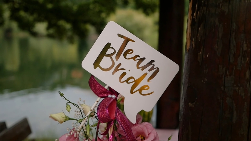 Tabla sa natpisom „Team Bride“ pričvršćena uz buket cveća, u prirodnom ambijentu pored vode