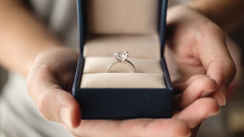 Osoba drži otvorenu tamnoplavu kutiju sa vereničkim prstenom od belog zlata i velikim kamenom u sredini
