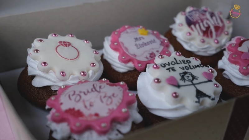 Kutija sa dekorisanim mafini­ma sa roze i belim ukrasima, natpisima „Bride to Be“ i malim crtežima vezanim za devojačko veče