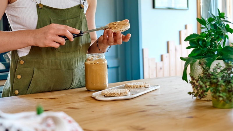 Osoba maže kikiriki puter na integralni tost u kuhinji, pored tegle kikiriki putera na stolu