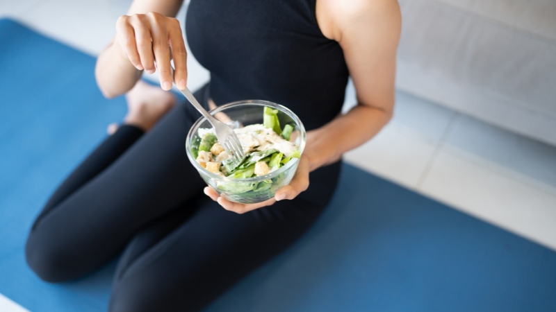 Osoba u sportskoj odeći sedi na prostirci i jede salatu