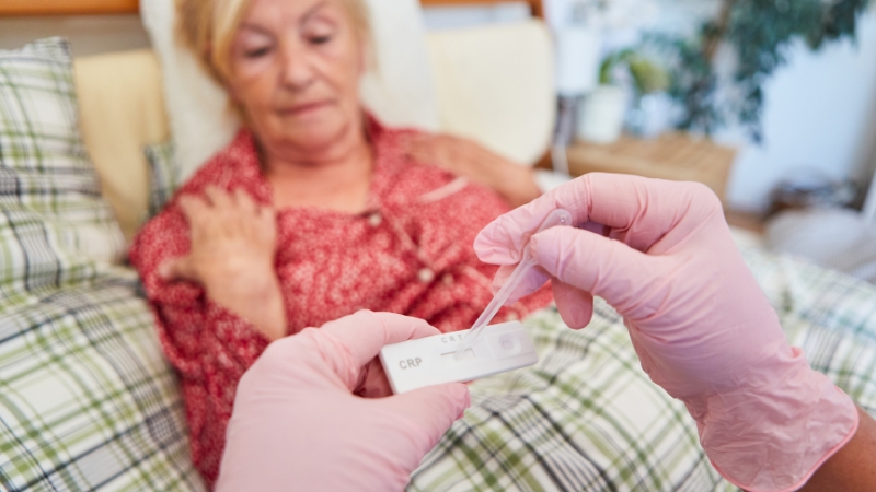Zdravstveni radnik u rukavicama radi brzi CRP test dok starija žena sedi u krevetu i drži se za rame